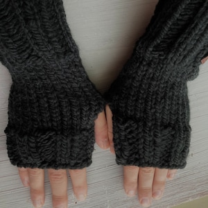 May include: A pair of black fingerless gloves made of knitted wool. The gloves are shown on a pair of hands with the fingers extended.