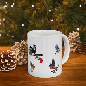 May include: White ceramic mug featuring a Christmas bird design. The mug depicts four birds, including a blue tit, a robin, a magpie, and a cardinal. The birds are shown in a festive setting with holly and a stocking.