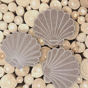May include: Three seashell-shaped coasters with a light brown color and white accent lines. The coasters are personalized with the names "Alyssa" and "Brandon". The coasters are displayed on a background of wooden logs.