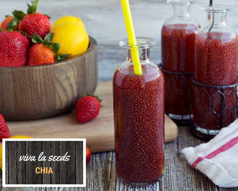 May include: A glass bottle filled with red chia seed pudding with a yellow straw sticking out of the top. The bottle is sitting on a wooden table next to a bowl of strawberries and a lemon. The text "viva la seeds CHIA" is on a wooden sign in the lower left corner of the image.