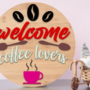 Op de afbeelding: Rond houten bord met de tekst "welcome coffee lovers" in rode en groene letters. Boven op het bord staan drie zwarte koffiebonen. Onderaan het bord staat een roze koffiekopje met stoom.