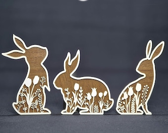 May include: Three wooden Easter bunny cutouts with floral designs. The bunnies are brown with white outlines and floral cutouts. The image includes the text "INSTANT DOWNLOAD" and "LASER READY FILES".