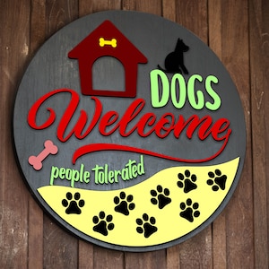 Puede incluir: Un letrero redondo de madera con las palabras "Dogs Welcome" en rojo y verde, y "people tolerated" en verde. El letrero presenta una caseta para perros, una silueta de perro, un hueso y huellas de patas.