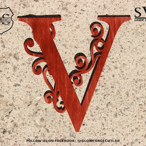 Puede incluir: Una letra V de madera roja con intrincados recortes de volutas. La letra está sobre un fondo beige texturizado. Archivos SVG listos para láser.