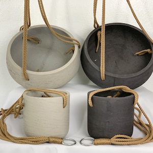 Puede incluir: Cuatro macetas colgantes, dos de color gris claro y dos de color gris oscuro. Cada maceta tiene un lazo de cuerda para colgar.