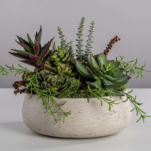 May include: A faux succulent arrangement in a white ceramic planter. The planter has a textured surface and the arrangement features a variety of green succulents.