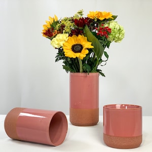 May include: Three pink ceramic vases with a textured, terracotta-colored base. One vase is upright, holding a bouquet of sunflowers and other colorful flowers. Another vase is lying on its side, and the third is a smaller, cylindrical vase.
