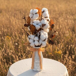 May include: A decorative arrangement of cotton bolls, acorns, and fall foliage in a repurposed bottle. The bottle is wrapped with a ribbon and has text that reads "HERITAGE SINCE 1913". The arrangement sits on a round table with a neutral tablecloth, set against a field.