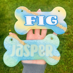 May include: Two bone-shaped pet tags. The top tag is blue, yellow, and white with the name "FIG" in white letters. The bottom tag is blue and green with the name "JASPER" in light green letters. Both tags have gold glitter accents.