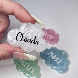 May include: Four cloud-shaped resin tags with glitter embedded in the resin. The tags are in different colours: white, pink, blue, and green. Each tag has a name written on it: "Clouds", "Rory", "Maya", and "Dexter".