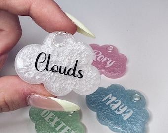 Può includere: Quattro etichette in resina a forma di nuvola con glitter incorporati nella resina. Le etichette sono di diversi colori: bianco, rosa, blu e verde. Ogni etichetta ha un nome scritto su di essa: "Clouds", "Rory", "Maya" e "Dexter".