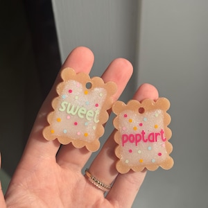Puede incluir: Dos llaveros con forma de Pop-Tart. Un llavero dice "sweet" en verde, y el otro dice "pop tart" en rosa. Ambos tienen un borde marrón claro y están decorados con chispas de colores.