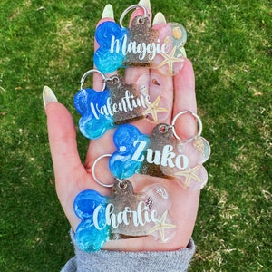 May include: Four bone-shaped dog tags made of blue and brown resin, each with a different name: Maggie, Valentine, Zuko, and Charlie. Each tag features a small seashell and a starfish embedded in the resin.