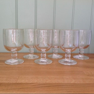 May include: A set of six clear glass goblets with etched cross designs. The goblets have a rounded bowl, a short stem, and a circular base. They are arranged on a wooden surface against a light green background.