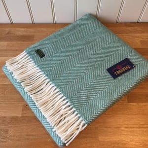 May include: A folded teal and white herringbone pattern blanket with white fringe. The blanket has a tag that reads "TWEEDMILL". The blanket is on a wooden surface.