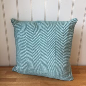 May include: A square, teal-colored pillow with a white herringbone pattern. The pillow is made of a textured fabric and has a soft, inviting appearance. The pillow is placed on a wooden surface against a white wall.