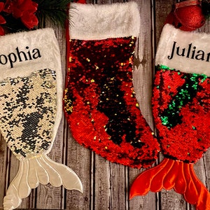 May include: Three Christmas stockings with mermaid tails. The stockings are red, white, and silver with sequins. The stockings are personalized with the names "Sophia", "Juliana", and a third name that is not visible in the image.