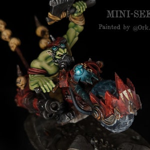 Puede incluir: Un Ork verde montando una motocicleta roja y negra con un diseño de calavera y huesos cruzados. La motocicleta tiene una rueda delantera grande con pinchos y una rueda trasera más pequeña con pinchos. El Ork sostiene un arma en su mano derecha y lleva una máscara de gas en la cara. El texto "MINI-SEKT" y "Pintado por @Ork.bot" es visible en la imagen.