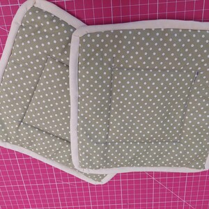 May include: Two square potholders featuring a sage green polka dot design with a cream border. The quilted potholders have a pocket on the front. These are designed for use in the kitchen.