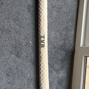 May include: A cream-coloured cricket bat grip with a diamond-patterned texture. The grip has the letters "TVR" printed in green. The grip is long and cylindrical, designed to wrap around the handle of a cricket bat.