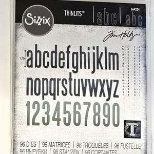Op de afbeelding: Een pakket Sizzix Thinlits stansmessen met het alfabet en cijfers. De letters en cijfers zijn in een schreefloze lettertype, ongeveer 2.2 cm hoog. Het pakket bevat 96 stansmessen en de tekst "Jim Holtz".