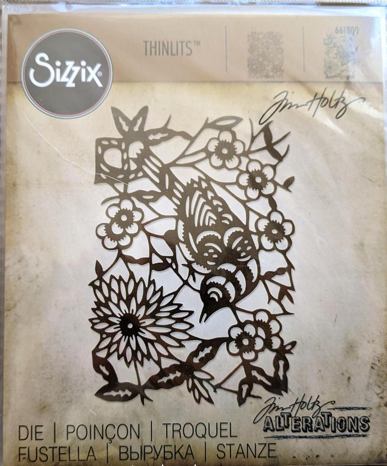 Op de afbeelding: Een Sizzix Thinlits stansvormenset met een gedetailleerd bloemen- en vogelontwerp. De uitgesneden afbeelding toont een vogel te midden van bloemen en bladeren. De verpakking bevat ook de tekst "Jim Holtz Alterations".