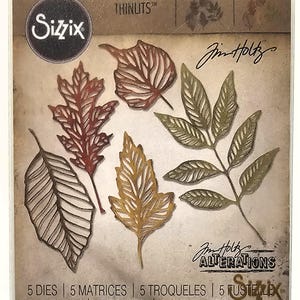 Op de afbeelding: Een pakket Sizzix Thinlits stansmessen met zes verschillende bladvormen in bruin, rood en goud. Het pakket bevat de tekst "Sizzix", "Thinlits", "Jim Holtz" en "Alterations". Het pakket bevat ook de tekst "5 DIES | 5 MATRICES | 5 TROQUELES | 5 FUSTELE | 5-ВЫРУБКИ | 5 STANZEN | 5 CORTANTES".