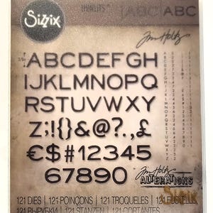 Op de afbeelding: Een Sizzix stansvormenset met het alfabet, cijfers en symbolen in een stencil-stijl lettertype. De set bevat 121 stansvormen en is ontworpen door Jim Holtz. De achtergrond heeft een vintage, verouderde papieruitstraling.
