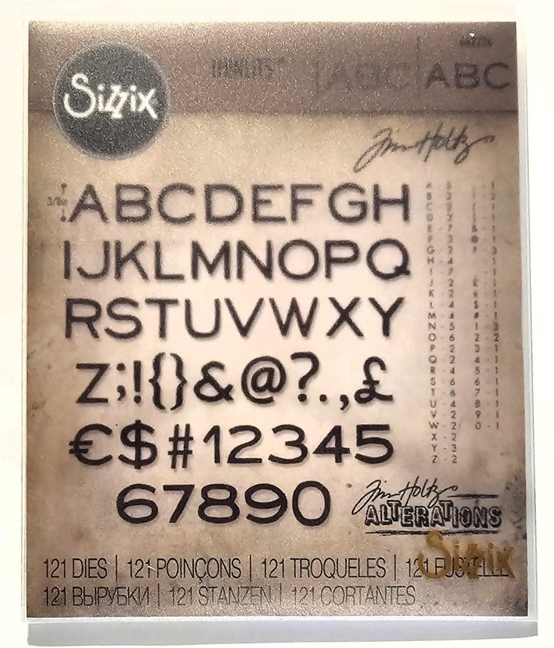 Op de afbeelding: Een Sizzix stansvormenset met het alfabet, cijfers en symbolen in een stencil-stijl lettertype. De set bevat 121 stansvormen en is ontworpen door Jim Holtz. De achtergrond heeft een vintage, verouderde papieruitstraling.