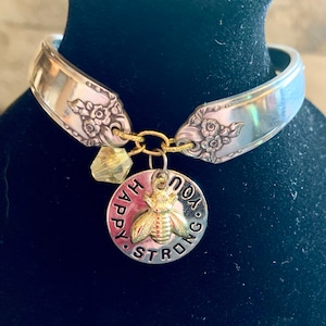 May include: A silver spoon bracelet with a gold charm that says "You are strong and happy" with a bee design.