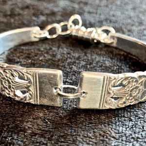 May include: Silver bracelet with floral and geometric patterns. The bracelet features a decorative design on the band and a clasp closure. The bracelet is approximately 7 inches in circumference.
