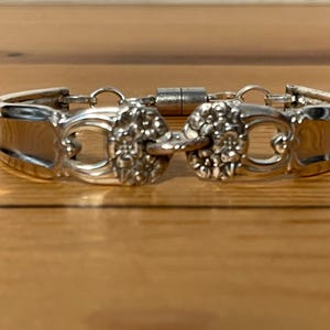 May include: A silver bracelet made from repurposed silverware. The bracelet features a floral design in the center and a magnetic clasp. The bracelet is displayed on a wooden surface.