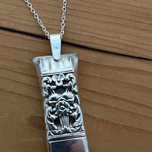 May include: A silver pendant necklace with a rectangular, ornate pendant. The pendant features a floral design with intricate details. The necklace chain is a delicate silver. The pendant is approximately 1.5 inches long.