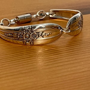 May include: A silver bracelet crafted from repurposed silverware. The bracelet features ornate floral and scrollwork details on the spoon bowls, connected by a chain and a magnetic clasp. The bracelet is displayed on a wooden surface.