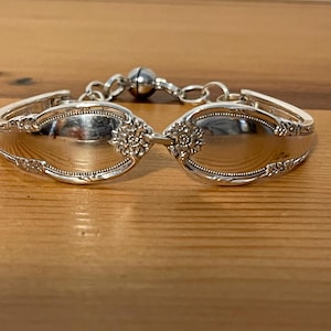 May include: A silver bracelet crafted from repurposed silverware. The bracelet features two spoon bowls connected by decorative floral accents and a magnetic clasp. The spoons have ornate detailing along the edges.