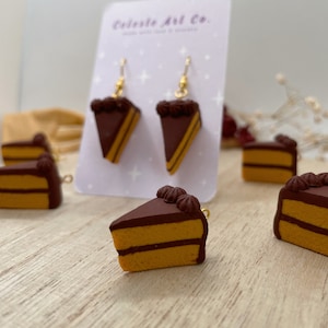 May include: A pair of gold dangle earrings with a chocolate cake slice charm. The cake slice is brown with yellow layers and white frosting. The earrings are displayed on a white card with the text "Celeste Art Co."
