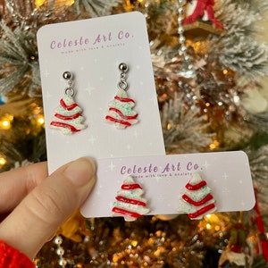 May include: A pair of silver stud earrings featuring white, red, and green candy cane Christmas tree designs. The earrings are displayed on a white card with the text "Celeste Art Co. made with love & anxiety".