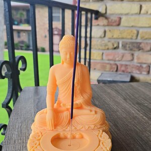 K&ouml;nnte beinhalten: Eine pfirsichfarbene Buddha-Statue mit einem R&auml;ucherst&auml;bchenhalter. Der Buddha befindet sich in einer sitzenden Meditationshaltung. Ein lila R&auml;ucherst&auml;bchen ist angez&uuml;ndet und in den Halter gelegt. Der Halter hat ein dekoratives, verziertes Design.