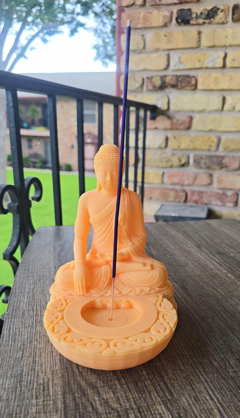 K&ouml;nnte beinhalten: Eine pfirsichfarbene Buddha-Statue mit einem R&auml;ucherst&auml;bchenhalter. Der Buddha befindet sich in einer sitzenden Meditationshaltung. Ein lila R&auml;ucherst&auml;bchen ist angez&uuml;ndet und in den Halter gelegt. Der Halter hat ein dekoratives, verziertes Design.