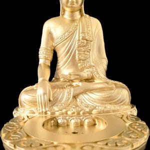 K&ouml;nnte beinhalten: Eine goldene Buddha-Statue in sitzender Meditationshaltung, vor schwarzem Hintergrund. Die Statue weist aufwendige Details in ihren Gew&auml;ndern und Verzierungen auf. Der Sockel hat ein dekoratives Design.
