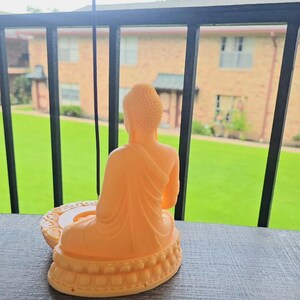 K&ouml;nnte beinhalten: Eine hellorangefarbene Buddha-Statue sitzt auf einem dekorativen Sockel. Die Statue befindet sich auf einer dunkelgrauen Oberfl&auml;che, mit einem gr&uuml;nen Rasen und einem Geb&auml;ude im Hintergrund. Ein R&auml;ucherst&auml;bchen befindet sich im Halter.