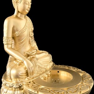 K&ouml;nnte beinhalten: Eine goldene Buddha-Statue in sitzender Meditationshaltung. Die Statue ist mit aufwendigen Designs versehen und steht auf einem dekorativen Sockel. Der Hintergrund ist schwarz.