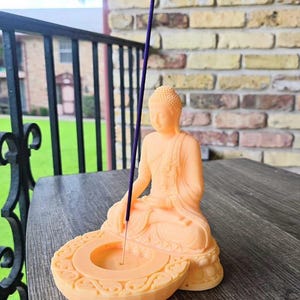 K&ouml;nnte beinhalten: Eine pfirsichfarbene Buddha-Statue mit eingebautem R&auml;ucherst&auml;bchenhalter. Die sitzende Figur h&auml;lt einen lila R&auml;ucherst&auml;bchen. Der Sockel der Statue hat ein dekoratives, kreisf&ouml;rmiges Design. Die Statue steht auf einer Holzoberfl&auml;che.