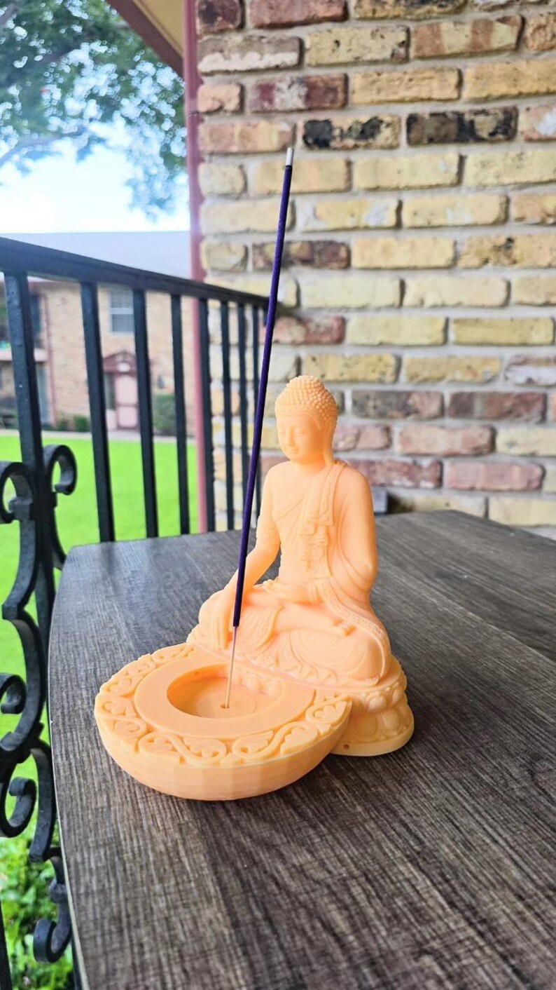 K&ouml;nnte beinhalten: Eine pfirsichfarbene Buddha-Statue mit eingebautem R&auml;ucherst&auml;bchenhalter. Die sitzende Figur h&auml;lt einen lila R&auml;ucherst&auml;bchen. Der Sockel der Statue hat ein dekoratives, kreisf&ouml;rmiges Design. Die Statue steht auf einer Holzoberfl&auml;che.