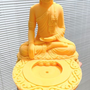 K&ouml;nnte beinhalten: Eine gelbe Buddha-Statue in sitzender Meditationshaltung. Die Statue weist komplizierte Details auf, darunter eine detaillierte Kopfbedeckung und drapierte Gew&auml;nder. Der Sockel hat ein verziertes, kreisf&ouml;rmiges Design mit einer zentralen &Ouml;ffnung.