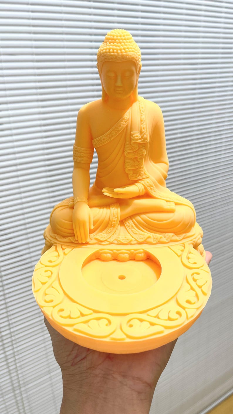 K&ouml;nnte beinhalten: Eine gelbe Buddha-Statue in sitzender Meditationshaltung. Die Statue weist komplizierte Details auf, darunter eine detaillierte Kopfbedeckung und drapierte Gew&auml;nder. Der Sockel hat ein verziertes, kreisf&ouml;rmiges Design mit einer zentralen &Ouml;ffnung.