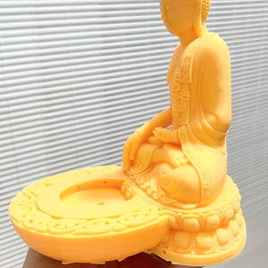 K&ouml;nnte beinhalten: Eine gelbe Buddha-Statue in sitzender Meditationshaltung. Die Statue steht auf einem runden Sockel mit dekorativem Rand. Der Kopf des Buddha hat ein strukturiertes Muster, und die Figur h&auml;lt die H&auml;nde in einer meditativen Geste.