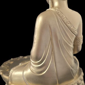 K&ouml;nnte beinhalten: Eine goldfarbene Buddha-Statue in sitzender Meditationshaltung. Die Statue weist auf der Robe und dem Lotus-Sockel aufwendige Details auf. Der Hinterkopf ist sichtbar, mit einem strukturierten Haardesign. Der Hintergrund ist schwarz.