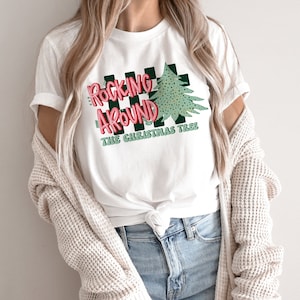 May include: White t-shirt with a green and black checkered design and the text "Rockin' Around The Christmas Tree" in red and pink letters.