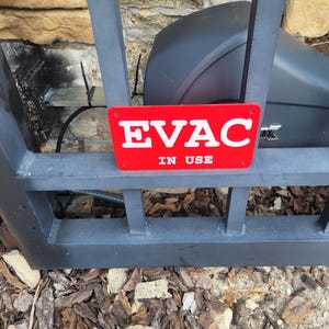 May include: A red rectangular sign with the white text "EVAC IN USE" is attached to a dark gray metal gate. The sign is in front of a black object and a stone wall.